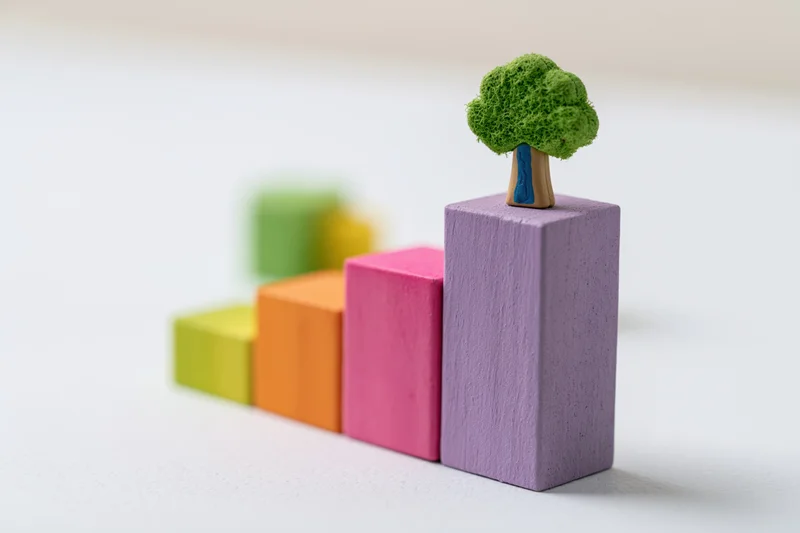 Colorful wooden blocks arranged in ascending bar chart with a small tree on the tallest block, symbolizing growth and sustainability