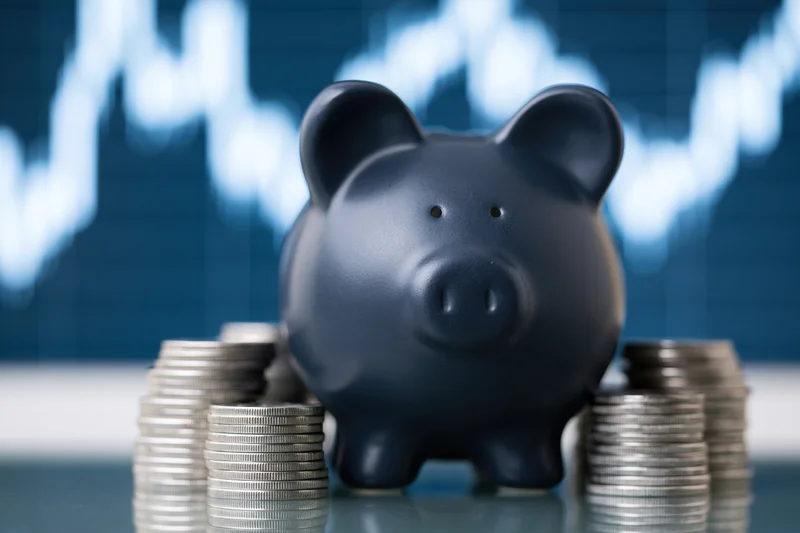 Dark navy ceramic piggy bank surrounded by stacks of coins on reflective surface with financial chart in background