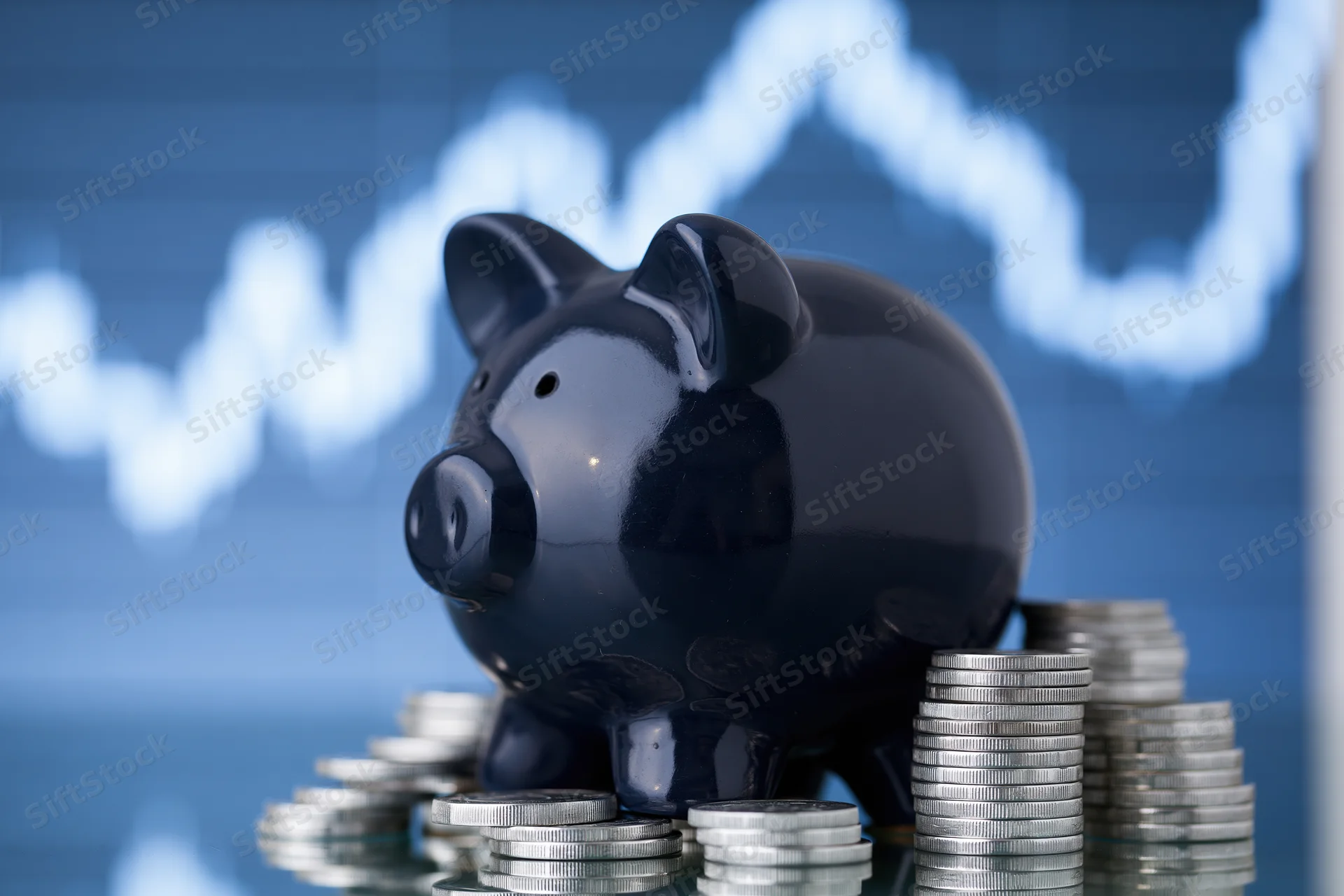 Dark navy ceramic piggy bank surrounded by stacks of coins on reflective surface with financial graph in background