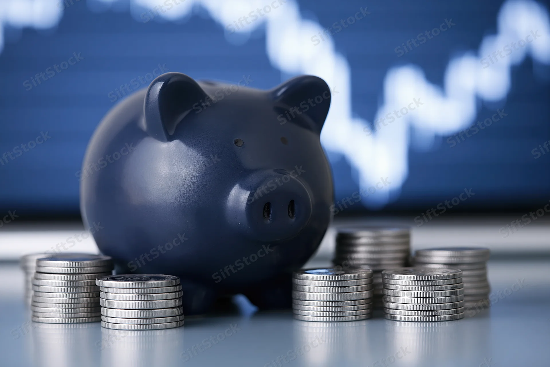 Dark navy ceramic piggy bank surrounded by stacks of coins on reflective surface with financial chart in background