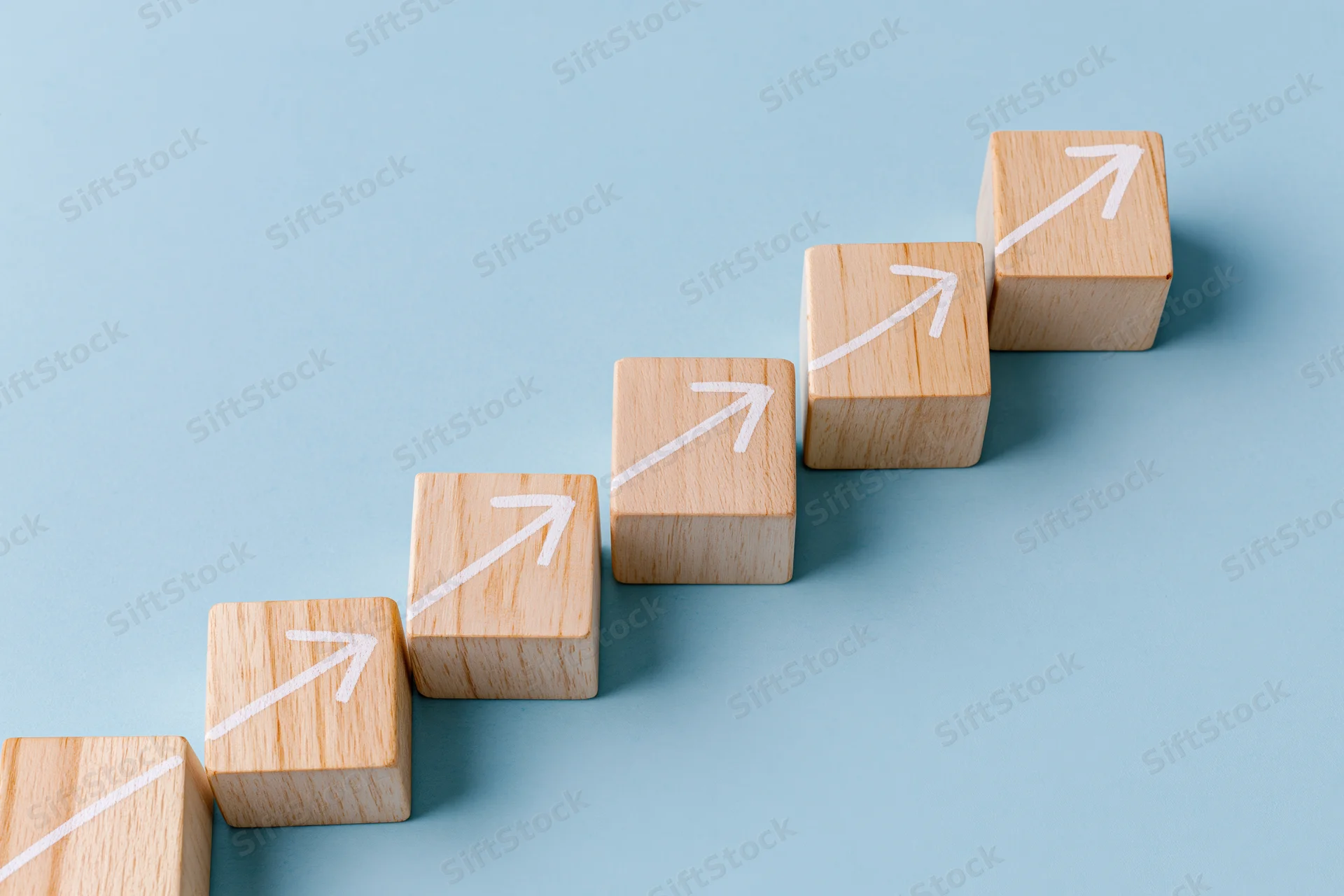 Wooden blocks with white arrows arranged in ascending staircase formation on light blue surface
