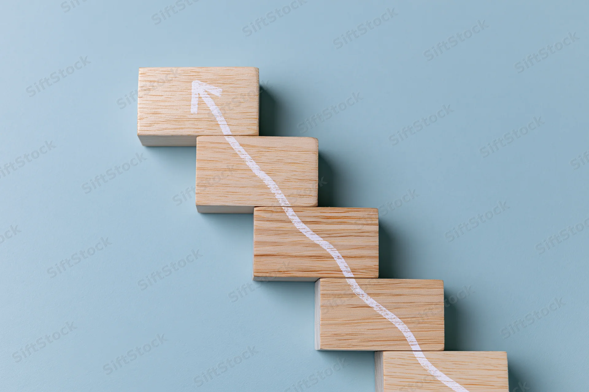 Wooden blocks arranged as ascending staircase with white arrow path on blue background