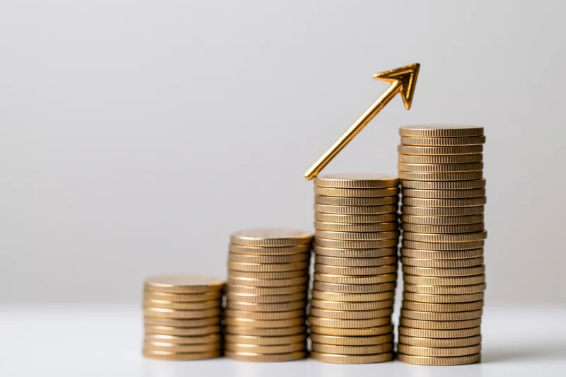 Stacks of gold coins increasing in height with a golden arrow pointing upward on a white surface