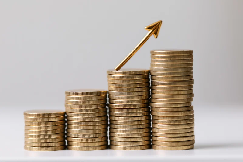 Stacks of gold coins increasing in height with a golden arrow pointing upward on a white surface