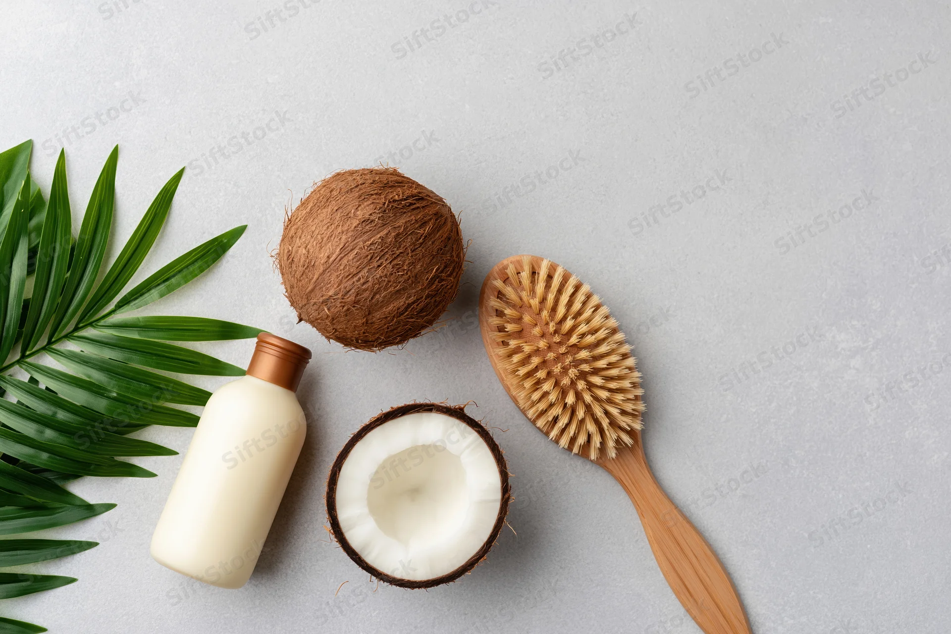 Flat lay of coconuts, a cream bottle, a wooden brush and palm leaves on a gray surface