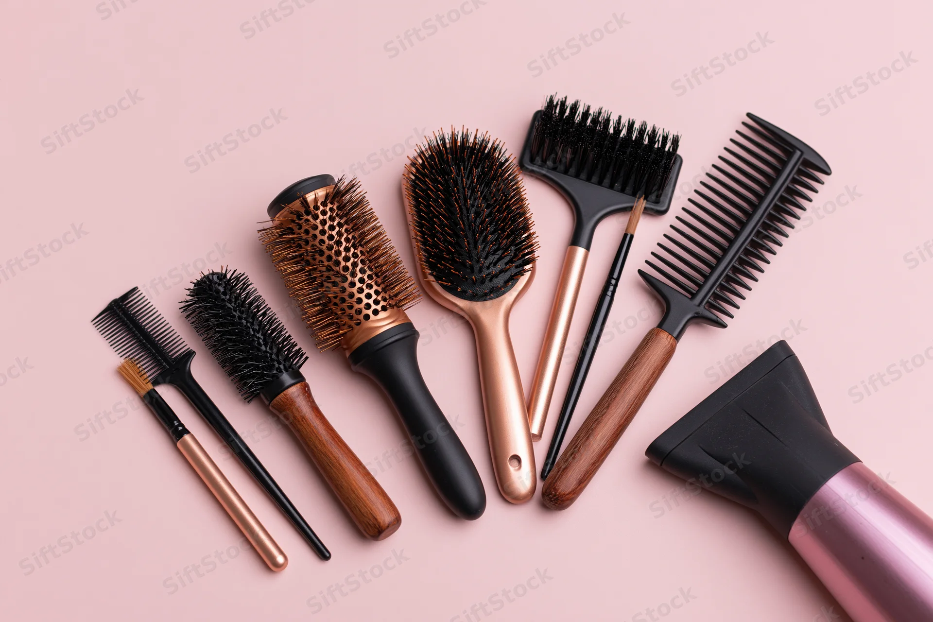 Assorted hairdressing tools including round barrel brushes and a hairdryer arranged in a flat lay on a pink background