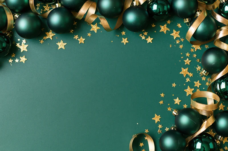 Dark green christmas ornaments and gold ribbon arranged in a frame on a matching green surface with scattered glitter stars and copyspace in the center