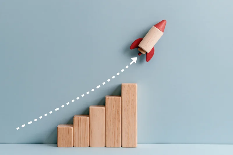 Wooden blocks arranged as a rising bar chart with a toy rocket launching upward along a dotted line