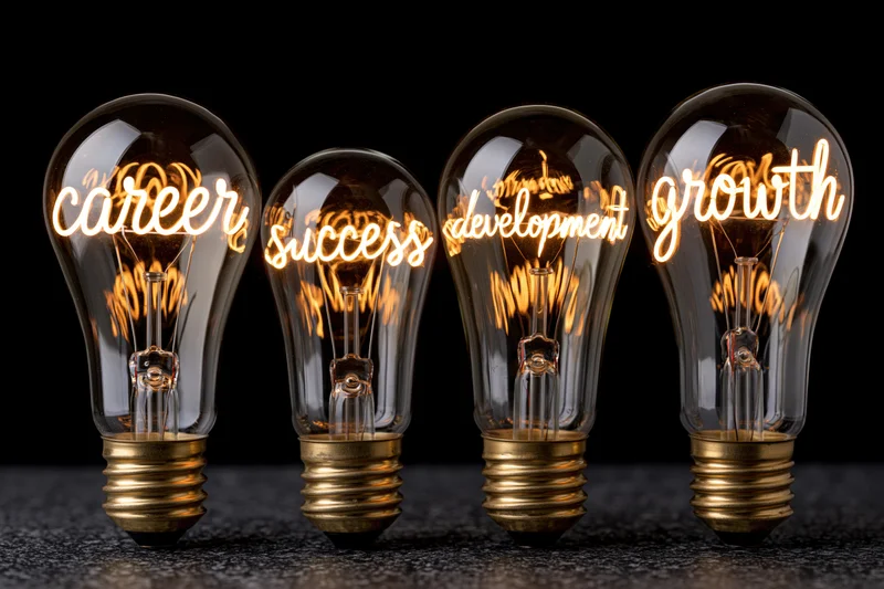 Four glowing incandescent lightbulbs in a row displaying the words career, success, development, and growth against a black background