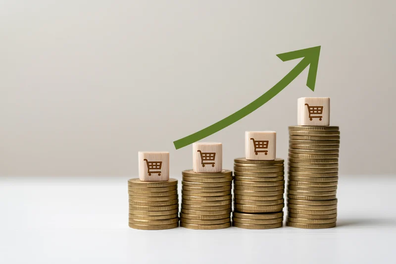Stacks of coins topped with shopping cart cubes and a green arrow trending upward on a white surface