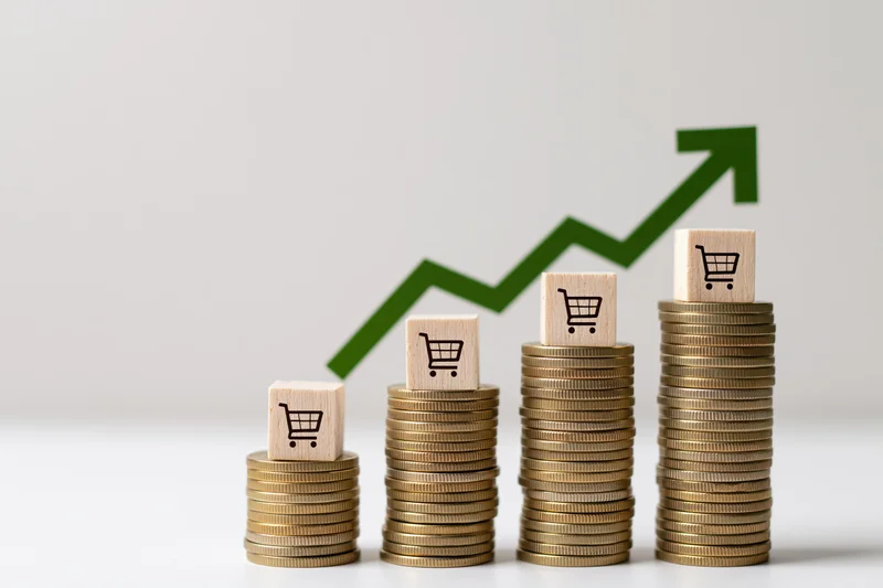 Stacks of coins topped with shopping cart icons arranged in ascending order with a green upward arrow in the background