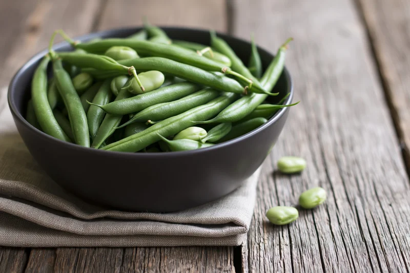 Fresh green beans in a dark ceramic bowl on a wooden table with a linen napkin