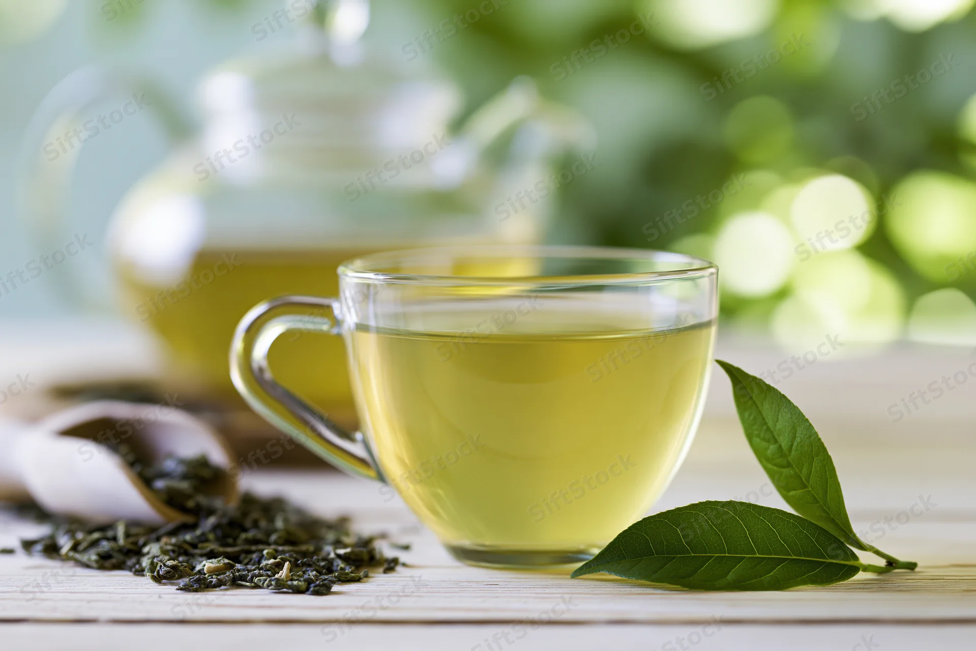 Glass cup of green tea on a pale wooden surface with fresh leaves and a teapot in soft focus