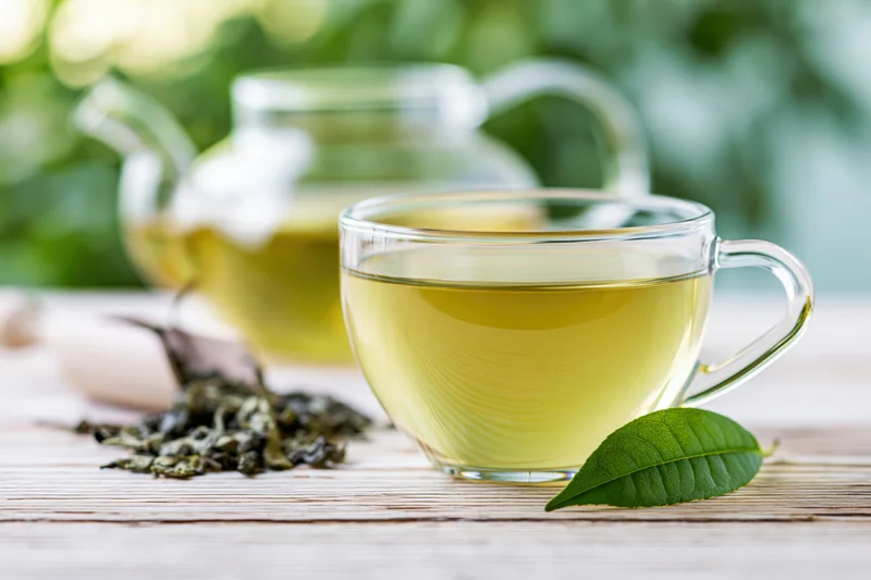 Glass cup of green tea on a pale wooden surface with dried leaves and a fresh leaf beside it