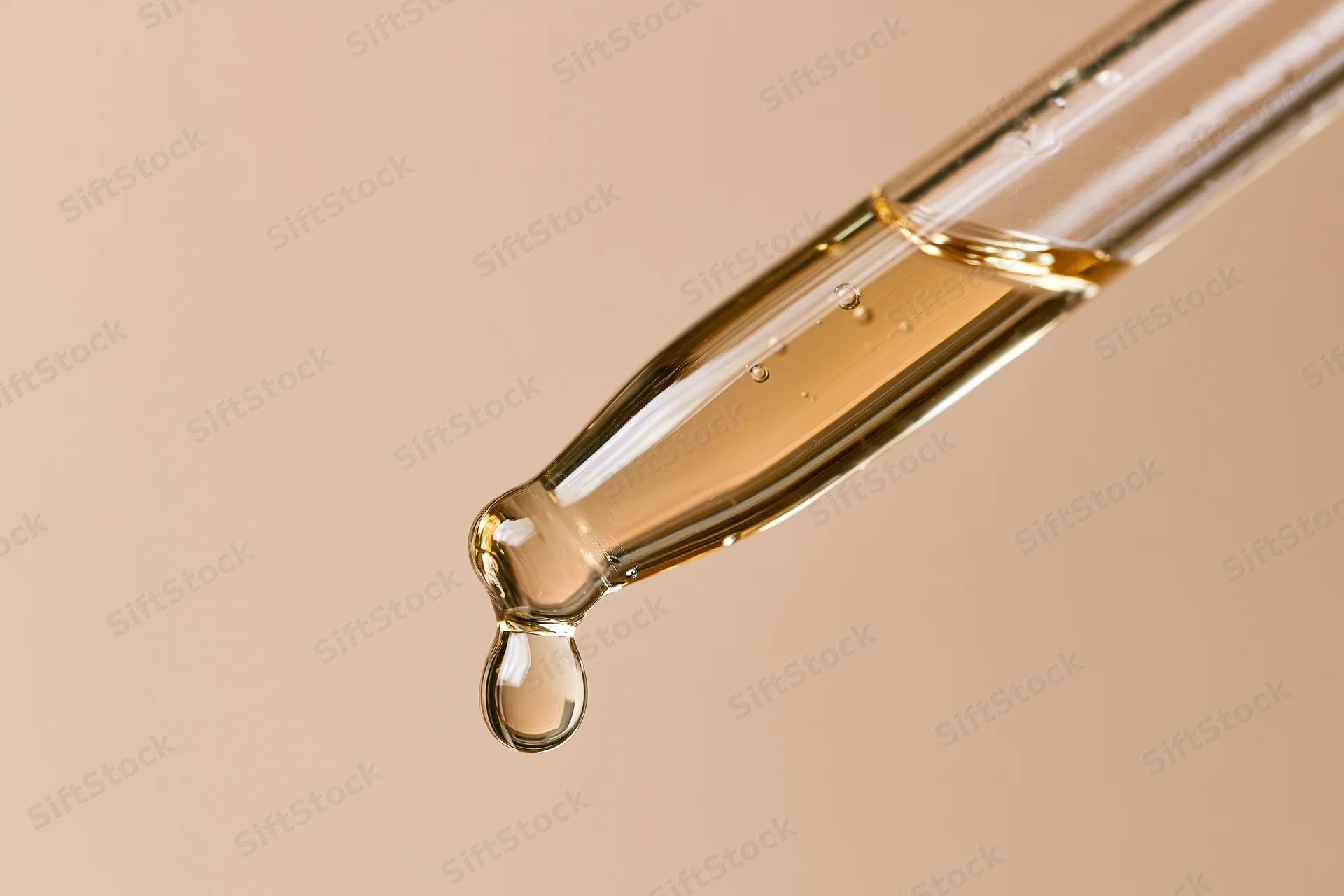 Glass pipette dropper with golden serum droplet against beige background
