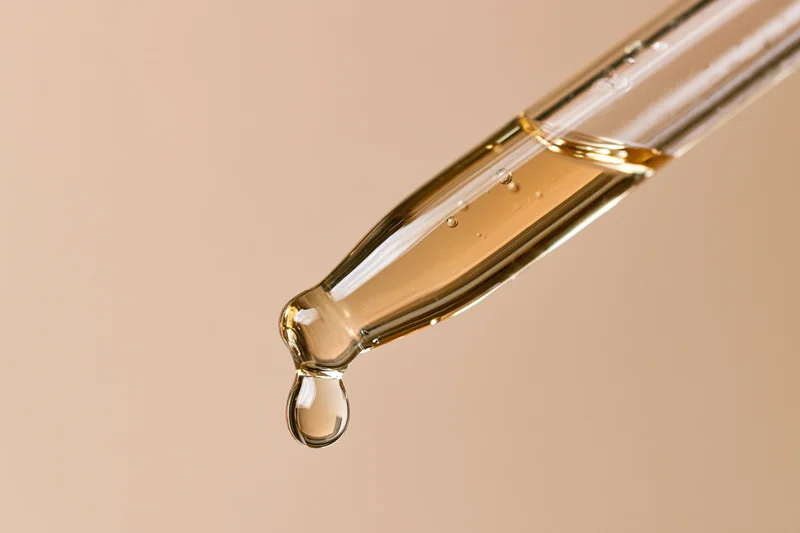 Glass pipette dropper with golden serum droplet against beige background