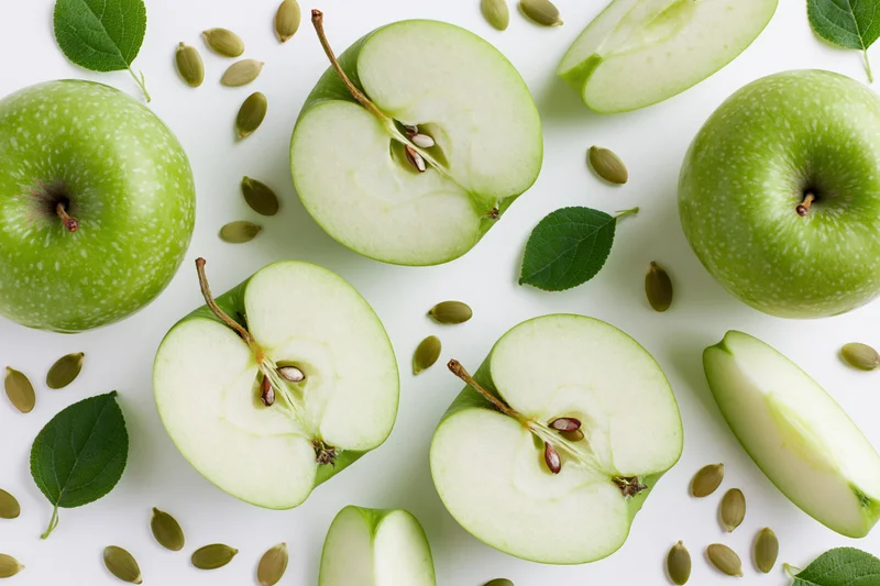 Green apples cut into halves and wedges arranged with leaves and seeds on white surface