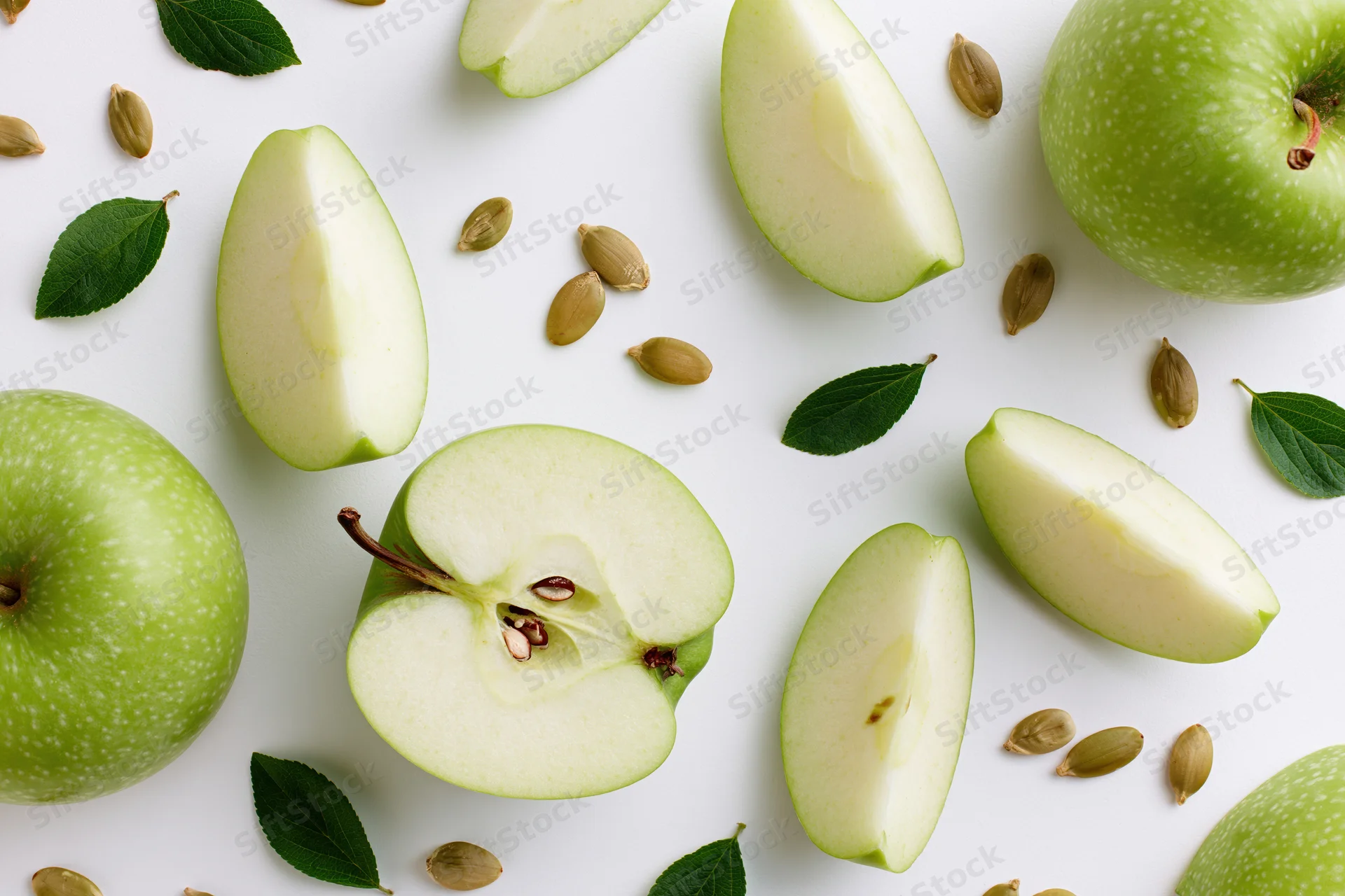 Green apples cut into halves and wedges arranged with leaves and seeds on white surface
