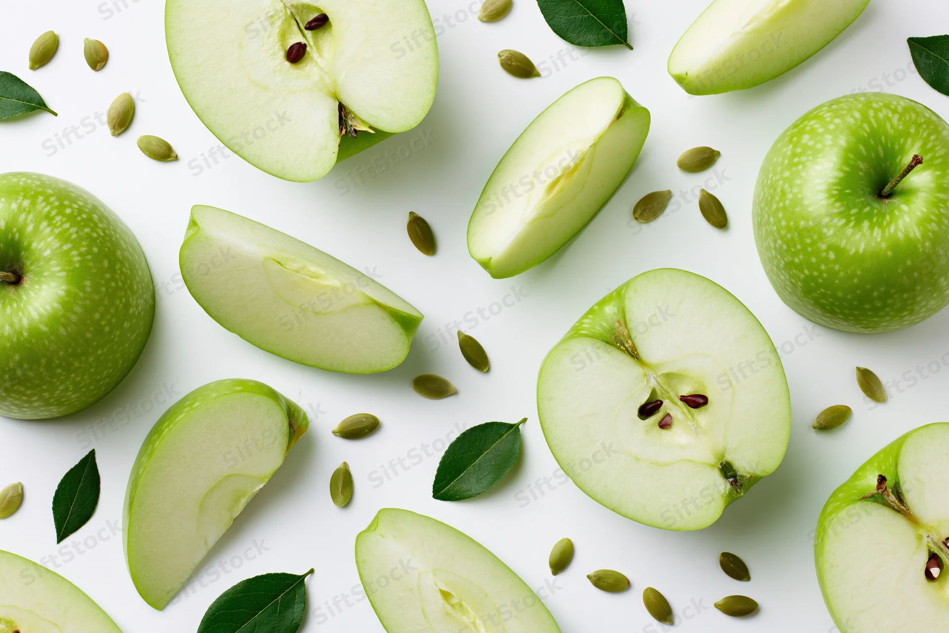 Green apples whole and sliced arranged with leaves and seeds on white surface
