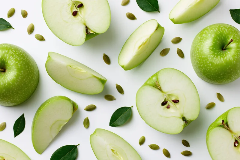 Green apples whole and sliced arranged with leaves and seeds on white surface