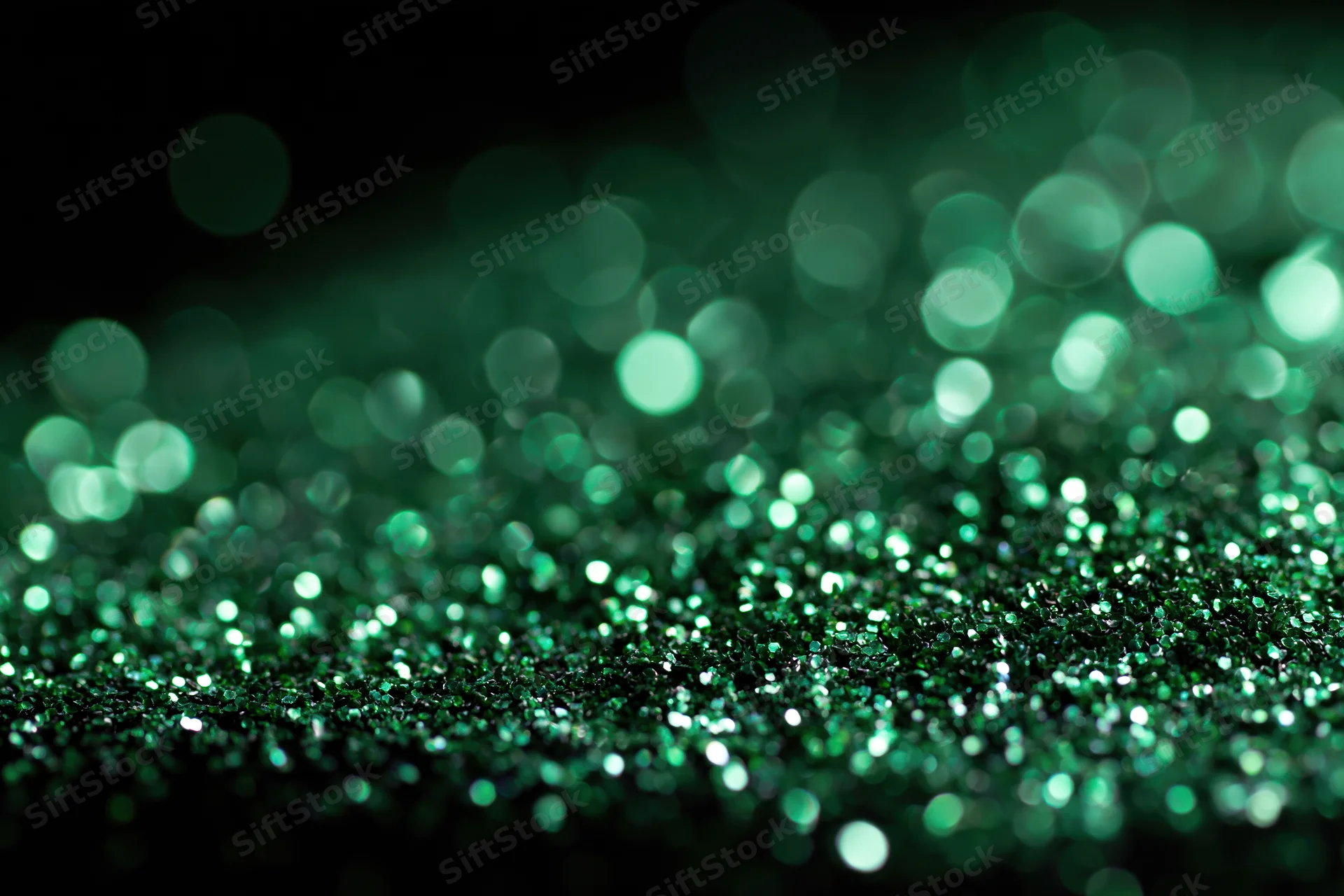 Green glitter particles scattered across a flat black surface with bokeh lights in the background