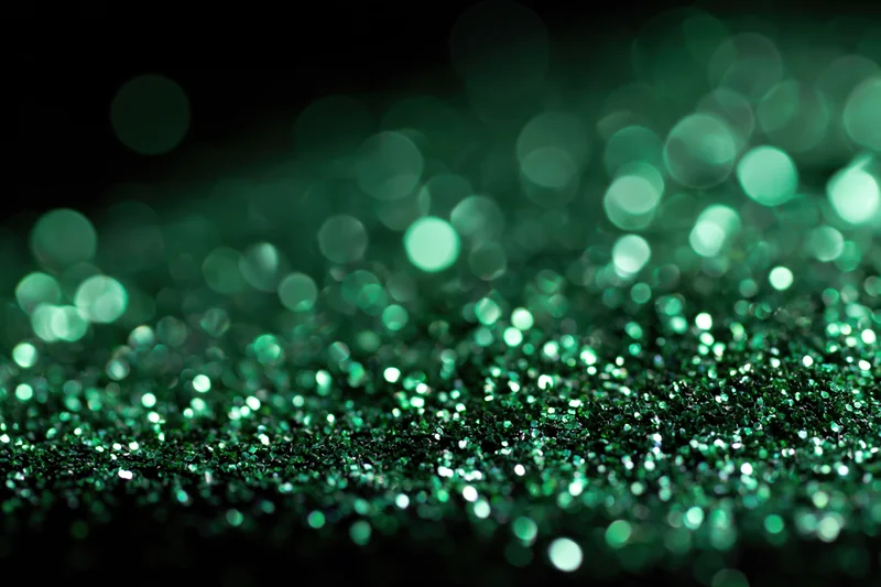 Green glitter particles scattered across a flat black surface with bokeh lights in the background