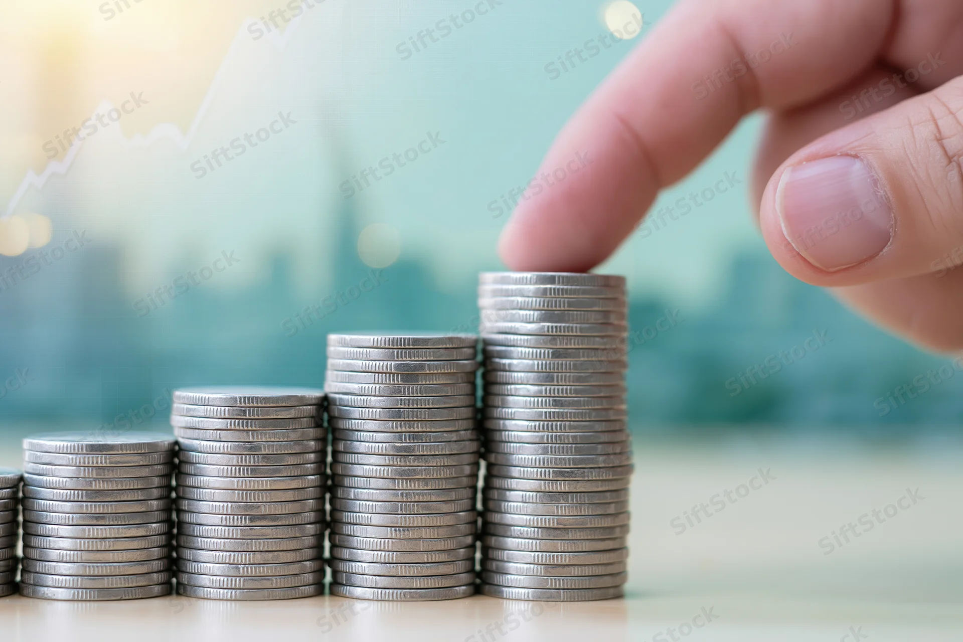 Hand placing a coin onto the tallest stack in a row of coins on a desk with a blurred financial graph in the background