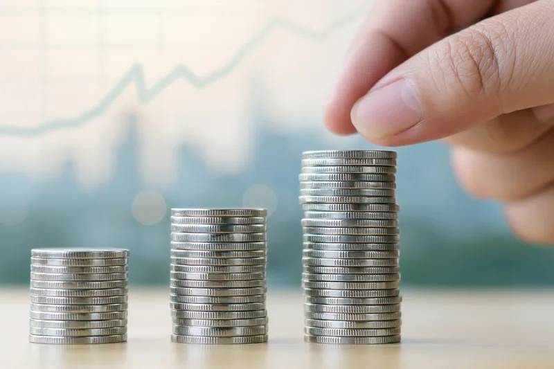 Hand placing a coin onto the tallest stack in a row of three on a table with a financial graph in the background