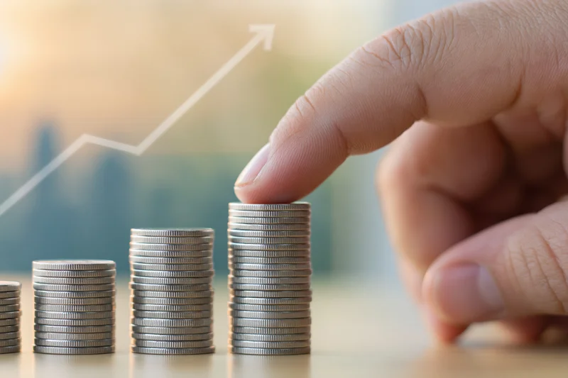 Hand placing a coin onto the tallest stack in a row of coins on a desk with a rising graph in the background