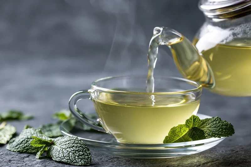 Hot green mint tea being poured from a glass teapot into a clear cup on a dark surface with fresh mint leaves around