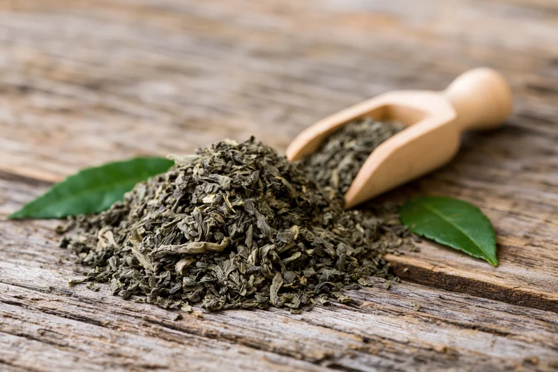 Pile of loose dried green tea leaves on weathered wooden surface with wooden scoop and fresh leaves