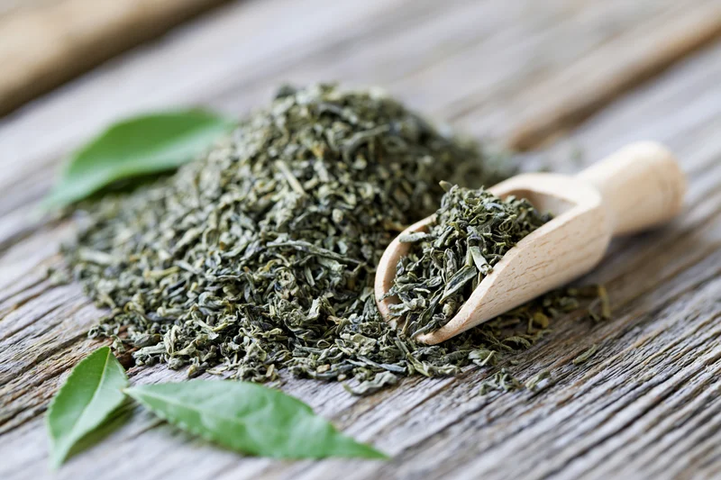 Loose dried green tea leaves piled on a weathered wooden surface with a wooden scoop and fresh leaves nearby