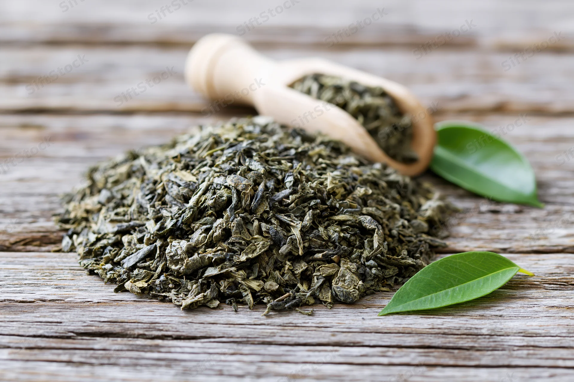 Pile of loose dried green tea leaves on weathered wooden surface with wooden scoop and fresh leaves