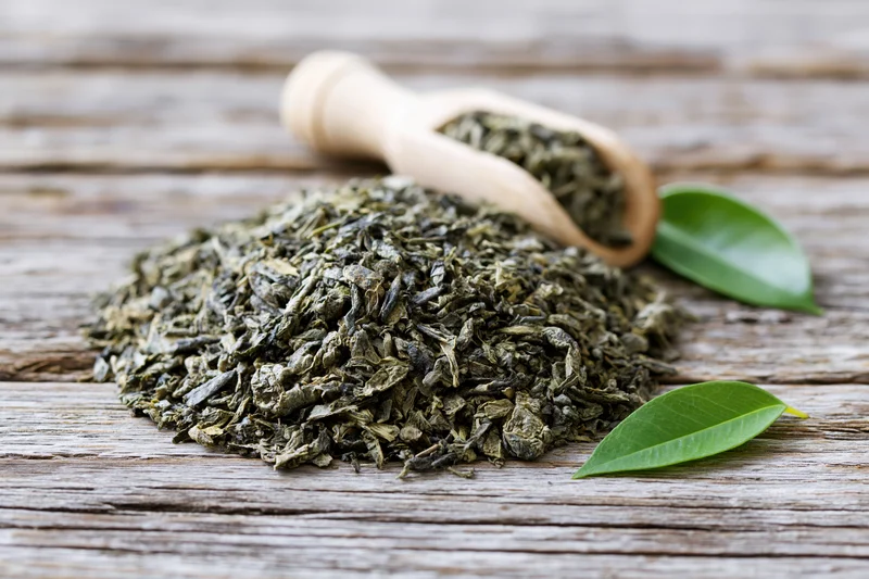 Pile of loose dried green tea leaves on weathered wooden surface with wooden scoop and fresh leaves