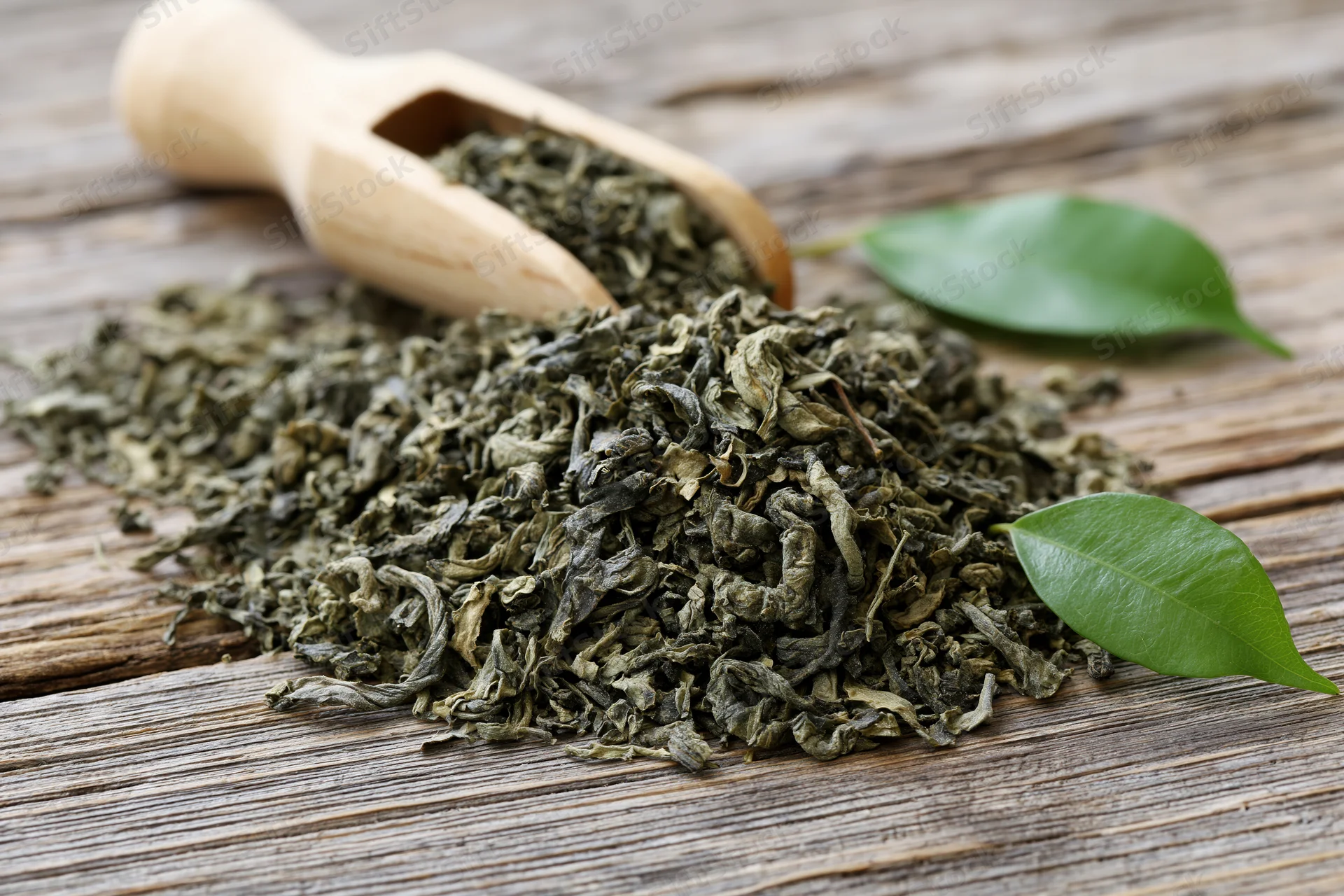 Loose dried green tea leaves piled on a weathered wooden surface with a bamboo scoop and fresh leaves nearby