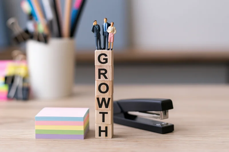 Miniature businesspeople standing on stacked wooden blocks spelling growth on an office desk