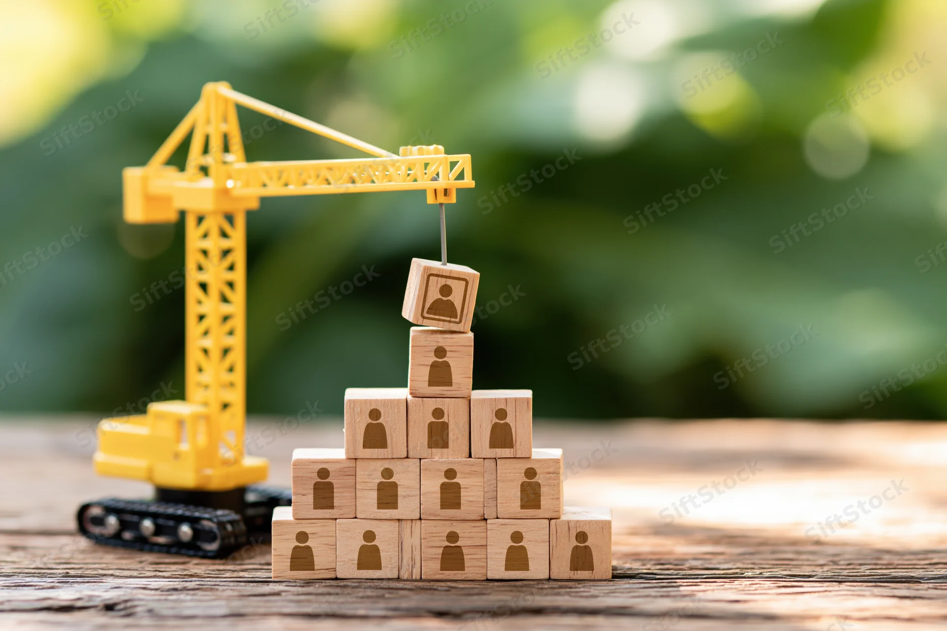 Miniature yellow toy crane lifting wooden blocks with human icons arranged in pyramid on wooden table outdoors