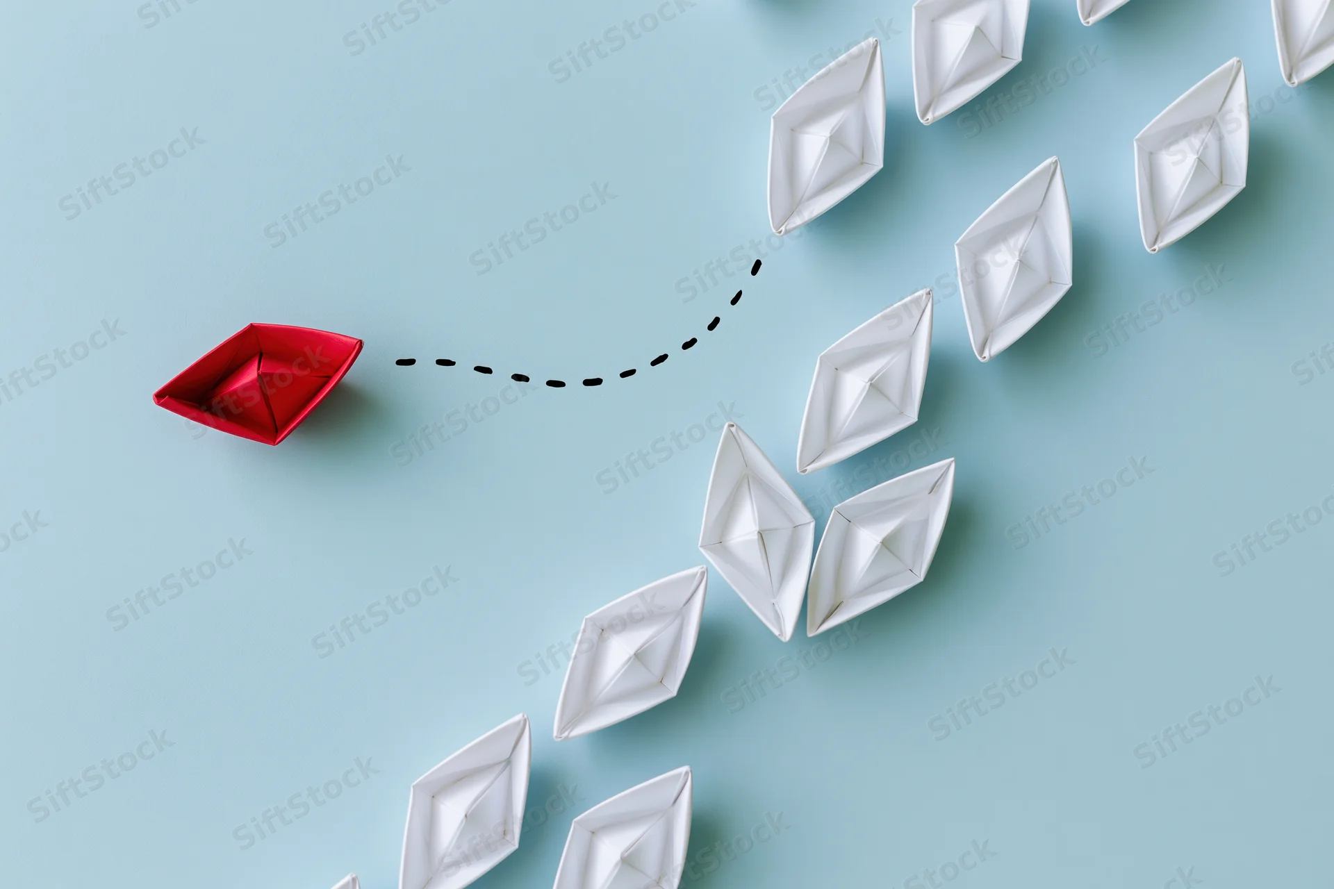 Red origami paper boat veering away from a group of white boats on blue surface