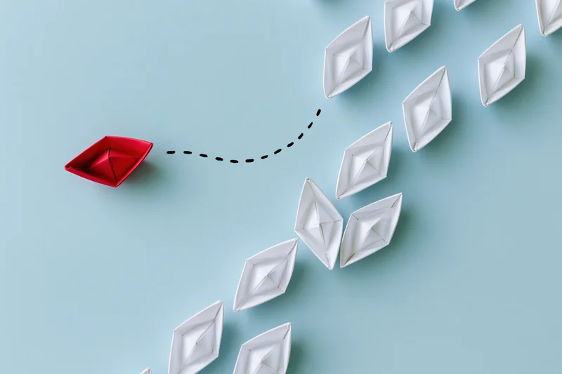 Red origami paper boat veering away from a group of white boats on blue surface