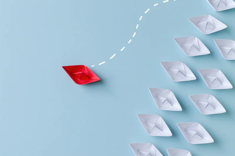 Red origami paper boat diverging from a group of white boats on a blue surface