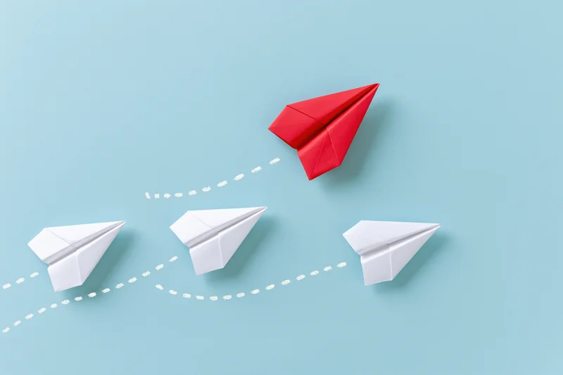 Red paper airplane flying upward while three white ones follow a curved path on a blue background