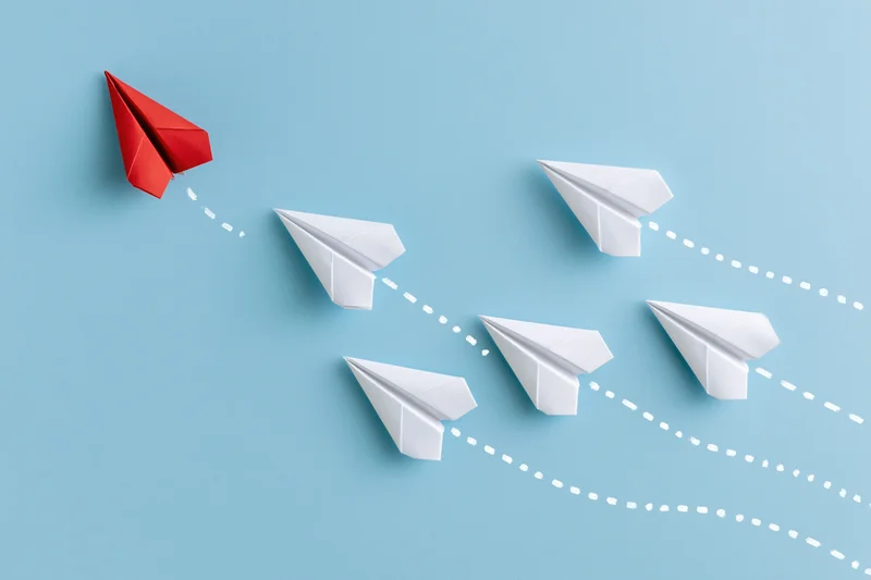 Red paper airplane leading white ones in flight against blue background