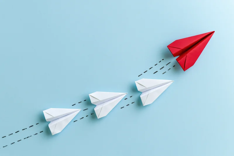 Red paper airplane leading white ones in flight against blue background