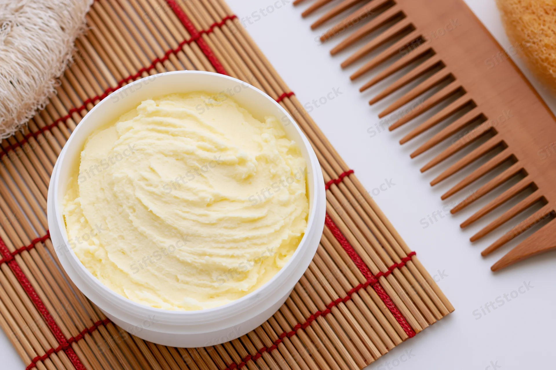 Open white plastic jar of pale yellow body butter cream on bamboo mat with wooden comb and sponge