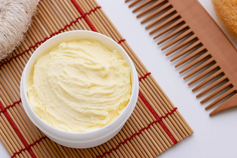 Open white plastic jar of pale yellow body butter cream on bamboo mat with wooden comb and sponge
