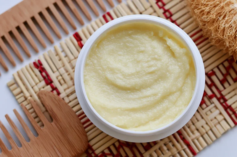Open white jar of pale yellow body butter cream on woven bamboo mat with wooden combs and brush