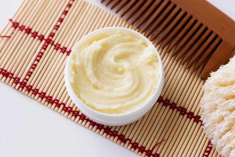 Open white jar of pale yellow body butter cream on bamboo mat with wooden comb and loofah