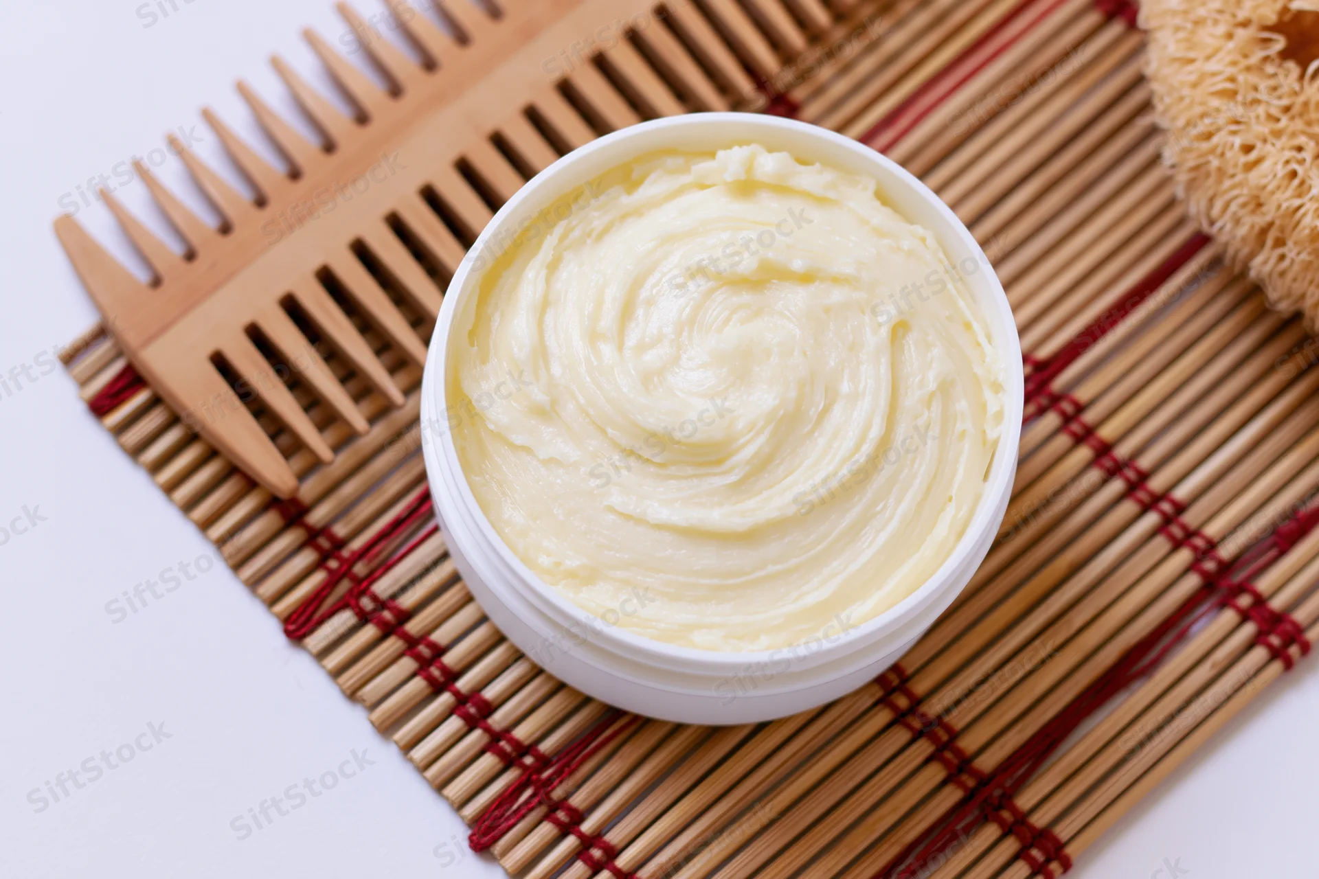Open white plastic jar of pale yellow body butter cream on bamboo mat with wooden comb and brush