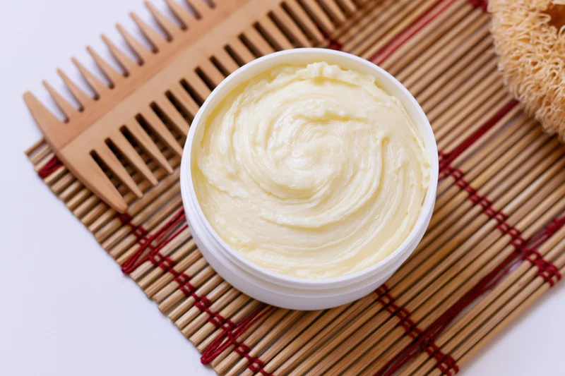 Open white plastic jar of pale yellow body butter cream on bamboo mat with wooden comb and brush