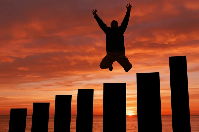 Silhouetted man jumping with arms raised above rising bar chart at sunset over ocean
